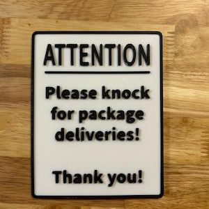 May include: A rectangular sign with a white background and black border. The sign reads "ATTENTION" above "Please knock for package deliveries!" and "Thank you!" in bold, black letters. The sign is on a wooden surface.