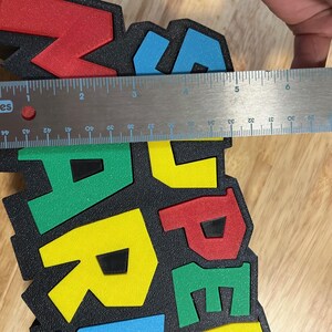 May include: A colorful foam sign with the word "MARIO" in red, yellow, green, and blue letters. The sign is approximately 5 inches wide. The letters are on a black background.