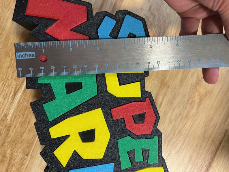 May include: A colorful foam sign with the word "MARIO" in red, yellow, green, and blue letters. The sign is approximately 5 inches wide. The letters are on a black background.