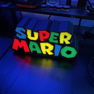 May include: A colorful Super Mario logo light. The letters are in red, yellow, blue, and green. The light is on a dark surface, with a blue hue. The letters are outlined in black.