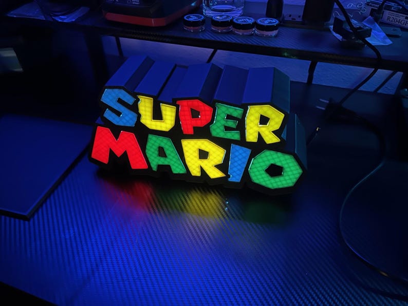 May include: A colorful Super Mario logo light. The letters are in red, yellow, blue, and green. The light is on a dark surface, with a blue hue. The letters are outlined in black.