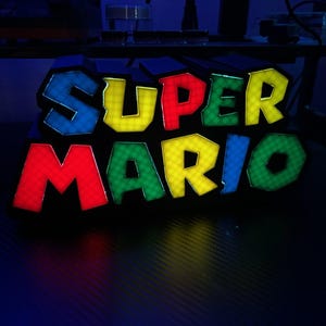 May include: A colorful, illuminated "SUPER MARIO" sign. The letters are outlined in blue and filled with yellow, red, green, and blue. The sign is likely a decorative item or a piece of gaming memorabilia, perfect for fans of the classic video game.
