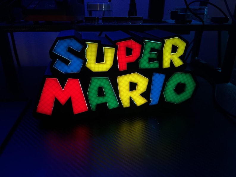 May include: A colorful, illuminated "SUPER MARIO" sign. The letters are outlined in blue and filled with yellow, red, green, and blue. The sign is likely a decorative item or a piece of gaming memorabilia, perfect for fans of the classic video game.