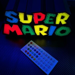 May include: A colorful LED sign displaying the words "SUPER MARIO" in bold, outlined letters. The letters are in red, yellow, green, and blue. A white remote control with multiple buttons is in the foreground. The sign is a gaming-themed product.