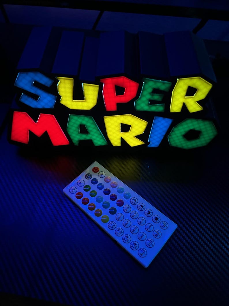 May include: A colorful LED sign displaying the words "SUPER MARIO" in bold, outlined letters. The letters are in red, yellow, green, and blue. A white remote control with multiple buttons is in the foreground. The sign is a gaming-themed product.
