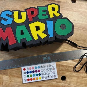 May include: A colorful "Super Mario" light-up sign with red, yellow, green, and blue letters. Includes a remote control and a USB cable. The sign is approximately 12 inches wide.