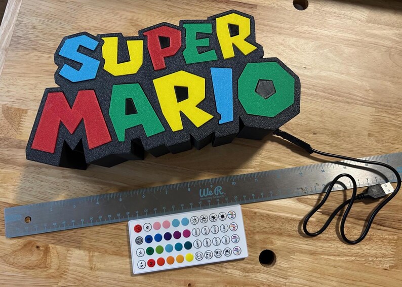 May include: A colorful "Super Mario" light-up sign with red, yellow, green, and blue letters. Includes a remote control and a USB cable. The sign is approximately 12 inches wide.