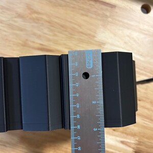 May include: A black, geometric object is measured with a metal ruler. The ruler shows measurements in inches. The object appears to be approximately 3 inches wide.