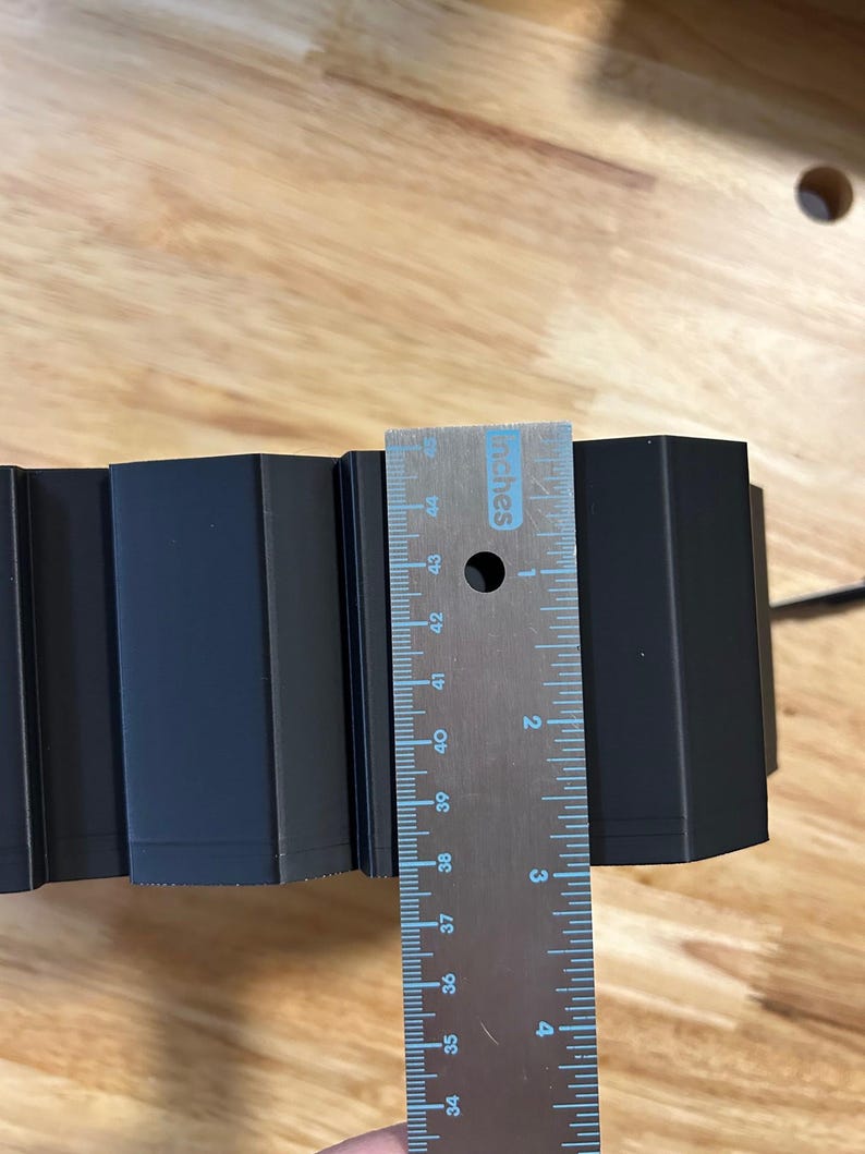 May include: A black, geometric object is measured with a metal ruler. The ruler shows measurements in inches. The object appears to be approximately 3 inches wide.