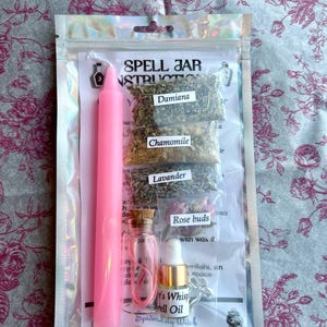 May include: A pink spell jar kit in a clear plastic bag. The kit includes a pink candle, small glass bottles, dried herbs labeled Damiana, Chamomile, and Lavender, and a bottle of spell oil. The text "Spell Jar Instructions" is visible.