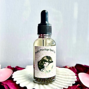 May include: A clear glass bottle with a black dropper top, labeled "Pretty Privilege Spell Oil." The label features an illustration of a woman. The bottle sits on a white, scalloped dish surrounded by red rose petals.