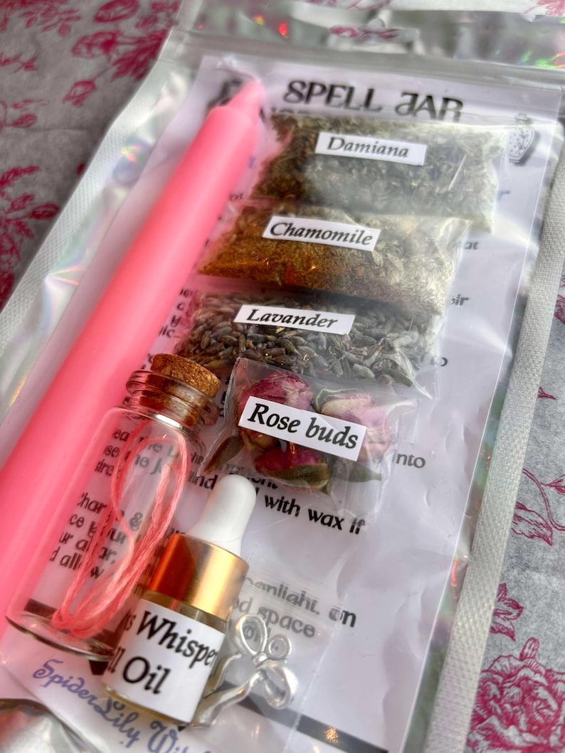 May include: A pink candle, small glass bottles, and packets of herbs and rose buds are inside a clear bag. The packets are labeled "Damiana", "Chamomile", "Lavender", and "Rose buds". A bottle of "Whisper Oil" is also included.