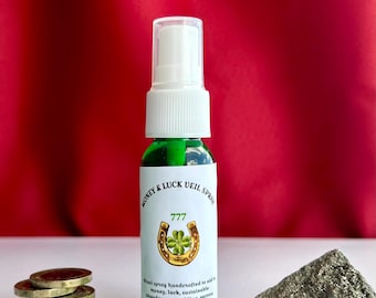 May include: A green glass spray bottle with a white cap and a label that reads "Money & Luck Veil Spray" with a horseshoe and four-leaf clover graphic. Beside it are a stack of coins and a piece of pyrite.