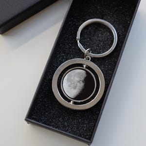 May include: A silver-colored keychain featuring a detailed image of the moon's phases. The circular pendant is encased in a double ring design and is presented in a black gift box. The moon image shows the phases of the moon.