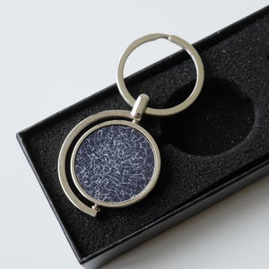 May include: A silver keyring with a circular pendant. The pendant has a navy blue background with a white, cracked design. The keyring is displayed in a black presentation box, suitable for gifting.