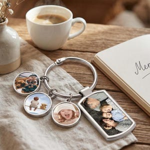 May include: A silver keyring with a large ring and several photo charms. The charms show colour photos of people. A white coffee cup and a notebook are in the background. The keyring rests on a beige cloth.