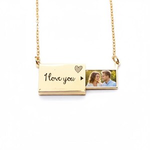 May include: A gold-colored rectangular locket necklace with a chain. The locket is engraved with "I love you" and a heart design. The locket opens to reveal a photograph of a couple. The necklace is displayed on a white background.