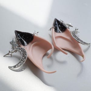 May include: A pair of elf ear prosthetics. The ears are a pale pink with black tips and silver-colored charms. The charms include crescent moons, stars, and starbursts. The ears are designed to be worn as costume accessories.