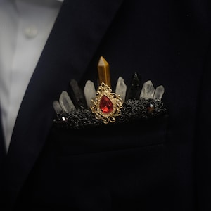 May include: A pocket square accessory featuring a black textured base adorned with an array of upright clear quartz, black tourmaline, and tiger's eye crystal points, centered with an ornate gold-tone filigree brooch holding a red teardrop gem.