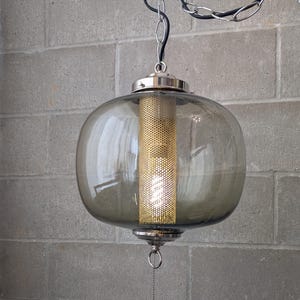 May include: A hanging pendant light with a smoky glass globe and a gold-toned, perforated metal cylinder at its center. The light fixture is suspended by a chain and features a pull chain with a black ball. The fixture is against a gray brick wall.