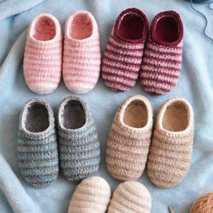 以下が含まれることがあります： ピンク、バーガンディ、ブルー、ベージュなど、さまざまな色の手編みスリッパのコレクション。明るいブルーの毛布の上に配置され、背景には毛糸とハサミが見えます。これらの快適なスリッパは、室内での使用に最適です。