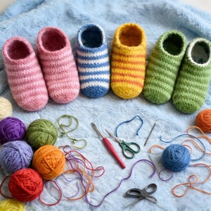 以下が含まれることがあります： ピンク、青、黄色、緑色の手編みスリッパが青い布の上に並べられています。ストライプのスリッパは、毛糸玉、かぎ針、ハサミ、針と一緒に配置されており、手作りのプロジェクトを示唆しています。