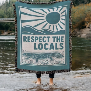 May include: Teal woven blanket with a graphic design featuring a sun, mountains, and a salamander. The text "RESPECT THE LOCALS" is prominently displayed. The blanket has a fringed edge and is held up by a person standing in water.