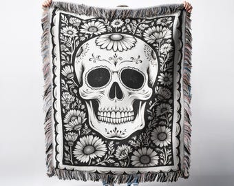 May include: Black and white woven throw blanket featuring a skull design surrounded by floral patterns. The blanket has a fringed edge with a mix of colors. The skull has detailed eye sockets, teeth, and decorative elements.