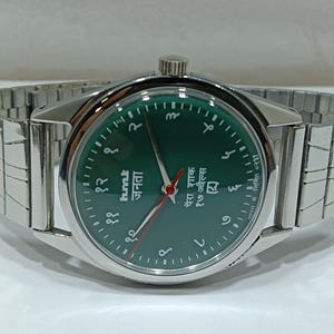 May include: A wristwatch with a green face and a silver-coloured strap. The watch face features Arabic numerals and text in a foreign language. The watch has a silver crown and a flexible metal strap.
