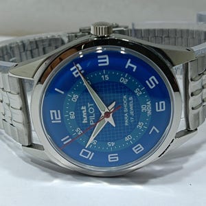May include: A silver wristwatch with a blue face. The watch face features white numerals and hands, with the text "hmt PILOT PARA SHOCK 17 JEWELS INDIA". The watch has a silver metal link strap.