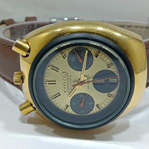 May include: A vintage Citizen Chronograph Automatic watch with a gold-tone case and a brown leather strap. The watch face is gold with black sub-dials and features the text "Citizen Chronograph Automatic 23 Jewels". The watch has gold-tone pushers and a crown.