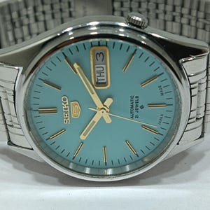 May include: A silver-toned Seiko 5 automatic wristwatch with a light blue dial. The watch features gold-colored hands and hour markers, a day-date display, and a stainless steel band. The watch face reads "SEIKO 5 AUTOMATIC 21 JEWELS MADE IN JAPAN".