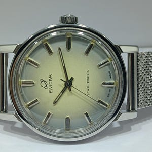 Può includere: Un orologio da polso Enicar Star Jewels color argento con cassa rotonda e quadrante chiaro sfumato. L'orologio presenta indici color argento, lancette sottili e un cinturino in maglia metallica. Sono visibili il testo ENICAR, un logo a forma di pianeta e SWISS MADE.