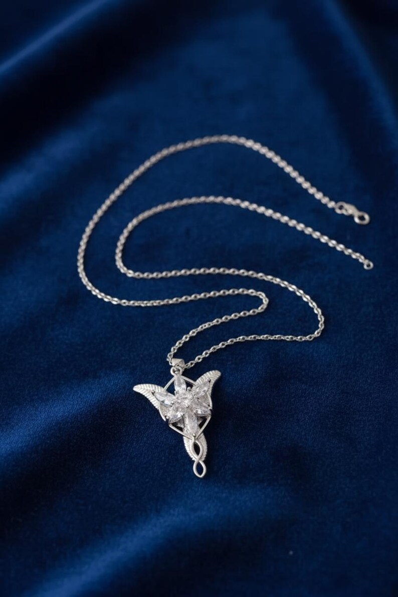 K&ouml;nnte beinhalten: Silberfarbene Halskette mit Anh&auml;nger, die ein stilisiertes Blumendesign mit klaren Kristallakzenten aufweist. Der Anh&auml;nger h&auml;ngt an einer zarten Kette vor einem tiefblauen Hintergrund. Geeignet f&uuml;r den t&auml;glichen Gebrauch oder besondere Anl&auml;sse.