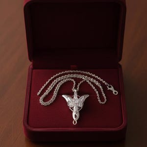 K&ouml;nnte beinhalten: Eine silberfarbene Halskette mit Anh&auml;nger und detailliertem Design, die in einer offenen, roten Samt-Schmuckschatulle liegt. Der Anh&auml;nger hat eine stilisierte Form mit aufwendigen Details und eine Kette. Die Schatulle steht auf einer Holzoberfl&auml;che.