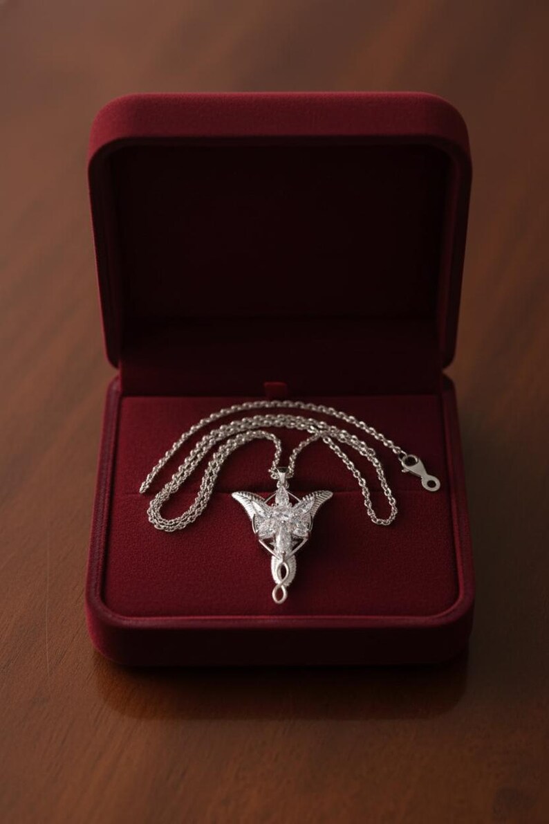 K&ouml;nnte beinhalten: Eine silberfarbene Halskette mit Anh&auml;nger und detailliertem Design, die in einer offenen, roten Samt-Schmuckschatulle liegt. Der Anh&auml;nger hat eine stilisierte Form mit aufwendigen Details und eine Kette. Die Schatulle steht auf einer Holzoberfl&auml;che.