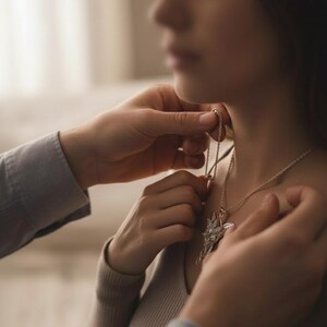 K&ouml;nnte beinhalten: Eine silberne Halskette mit einem sternf&ouml;rmigen Anh&auml;nger wird einer Person um den Hals gelegt. Der Anh&auml;nger hat ein zentrales Design mit strahlenf&ouml;rmigen Elementen. Die Person tr&auml;gt einen hellfarbigen Pullover.