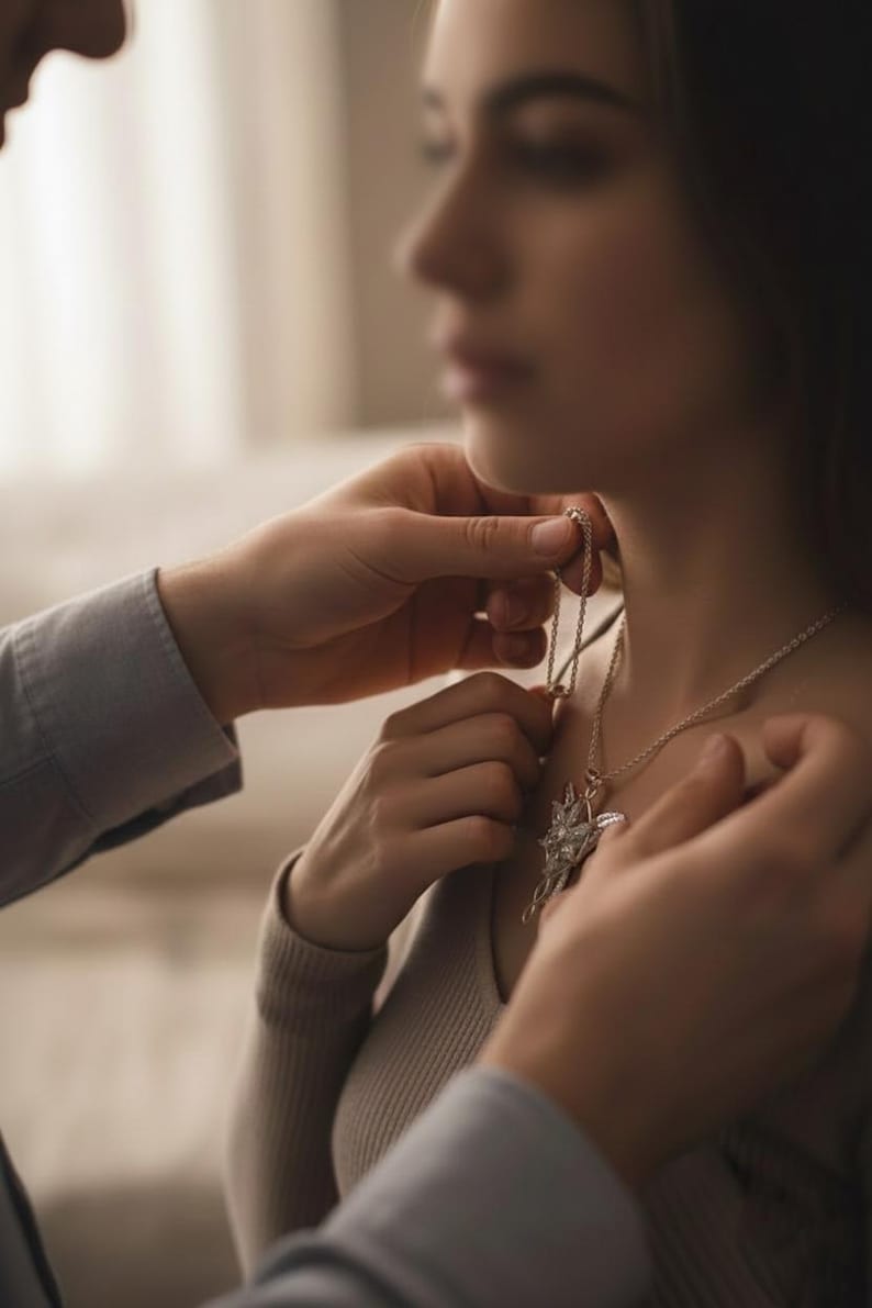 K&ouml;nnte beinhalten: Eine silberne Halskette mit einem sternf&ouml;rmigen Anh&auml;nger wird einer Person um den Hals gelegt. Der Anh&auml;nger hat ein zentrales Design mit strahlenf&ouml;rmigen Elementen. Die Person tr&auml;gt einen hellfarbigen Pullover.
