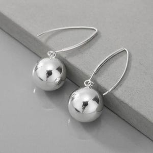 May include: A pair of silver-toned drop earrings featuring polished, spherical ball charms suspended from curved, open-loop ear wires. The smooth, reflective metal surfaces of the spheres catch the light, creating a bright, metallic appearance.