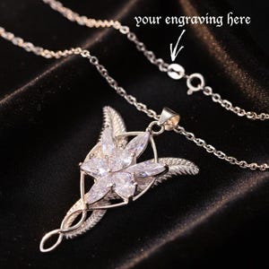 K&ouml;nnte beinhalten: Eine silberfarbene Halskette mit einem Anh&auml;nger in floralem Design mit klaren Edelsteinen. Die Kette ist filigran, und der Anh&auml;nger hat ein detailliertes, stilisiertes Blattdesign. Der Text auf dem Bild lautet "your engraving here".