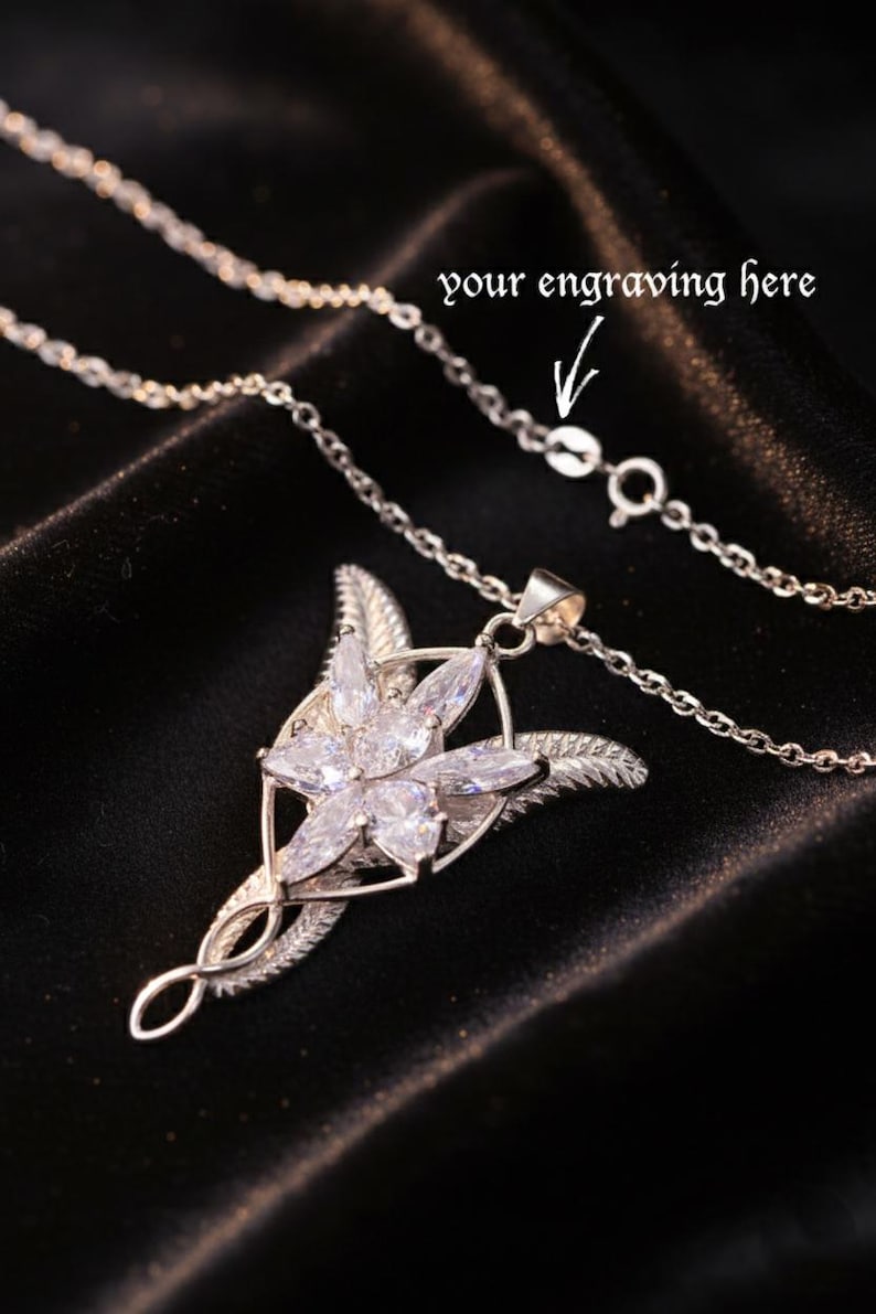 K&ouml;nnte beinhalten: Eine silberfarbene Halskette mit einem Anh&auml;nger in floralem Design mit klaren Edelsteinen. Die Kette ist filigran, und der Anh&auml;nger hat ein detailliertes, stilisiertes Blattdesign. Der Text auf dem Bild lautet "your engraving here".