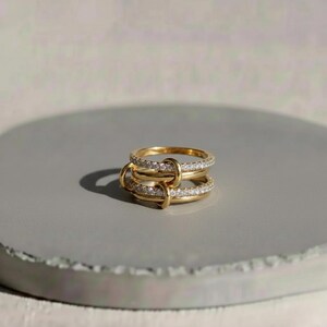 May include: A gold-colored ring with three bands, each encrusted with small, clear gemstones. The bands are connected by gold loops, creating a unique design. The ring is displayed on a gray surface, showcasing its intricate details and elegant style.