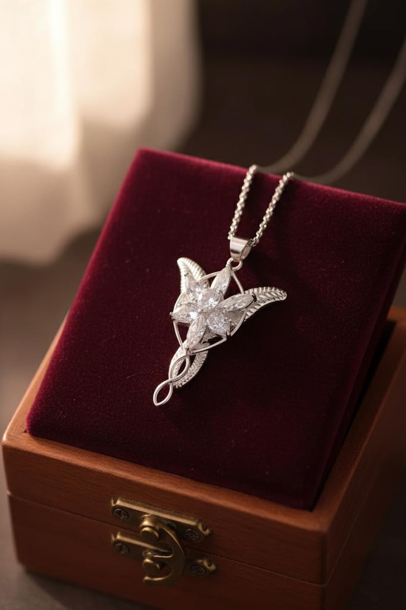 K&ouml;nnte beinhalten: Eine silberfarbene Halskette mit Anh&auml;nger, die ein sternf&ouml;rmiges Design mit klaren, funkelnden Edelsteinen aufweist. Der Anh&auml;nger h&auml;ngt an einer zarten Kette und liegt auf einer bordeauxroten Samtoberfl&auml;che in einer Holzbox. Die Halskette ist ein Schmuckst&uuml;ck.