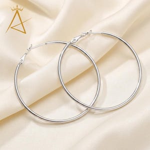 May include: A pair of large, silver hoop earrings. The earrings are round and have a simple, polished design. They are displayed on a cream-colored, satin-like fabric. The earrings are a classic jewelry item.