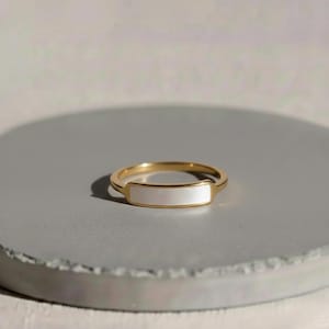 May include: A gold-colored ring with a rectangular, white mother-of-pearl inlay. The ring is set on a gray surface, showcasing its polished finish. The band is thin and delicate, highlighting the elegant design of the jewelry.