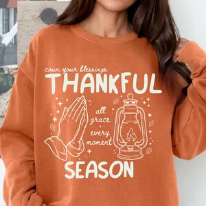 Puede incluir: Una sudadera de cuello redondo color terracota con gráficos serigrafiados en blanco. El diseño incluye manos en oración, una linterna y el texto: count your blessings, THANKFUL, all grace, every moment, SEASON.