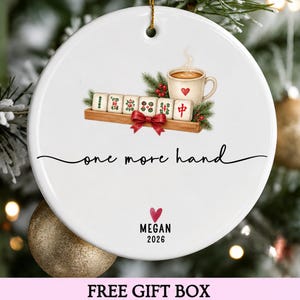 Puede incluir: Un adorno redondo de cerámica blanca con un gráfico de fichas de mahjong en un soporte de madera, una taza de café y un lazo rojo. El texto dice one more hand, MEGAN y 2026, con un pequeño icono de corazón rojo bajo el texto.
