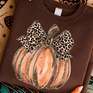 Puede incluir: Una sudadera de cuello redondo en color marrón chocolate oscuro con un gráfico central de una calabaza con un lazo de estampado de leopardo. La calabaza tiene una estética pintada en tonos rosa claro y naranja, con detalles en dorado brillante.