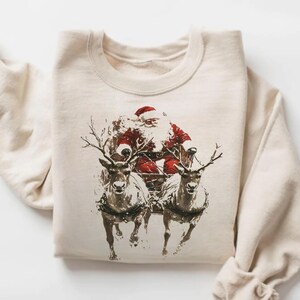 Puede incluir: Una sudadera de cuello redondo color arena con un estampado gráfico de Santa Claus en un trineo tirado por dos renos. La ilustración utiliza tonos marrón oscuro y rojo para representar las figuras con un estilo detallado y tipo boceto.
