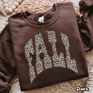 Puede incluir: Una sudadera de cuello redondo en color marrón chocolate oscuro con la palabra FALL en letras grandes de estilo bloque. Las letras están rellenas con un estampado de leopardo en tonos beige, negro y marrón y tienen un borde claro.